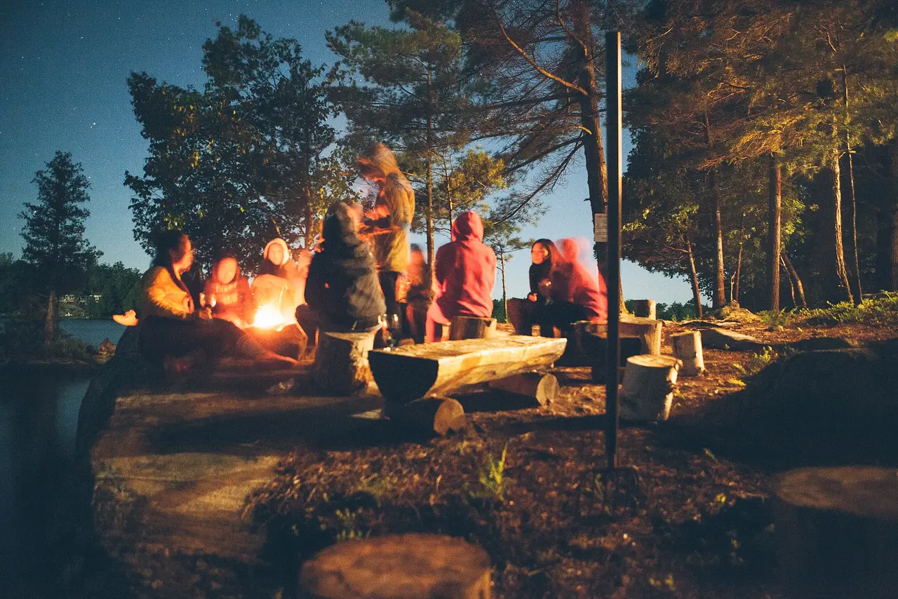 Family and friends telling scary stories while sitting on logs around a campfire in the woods.
